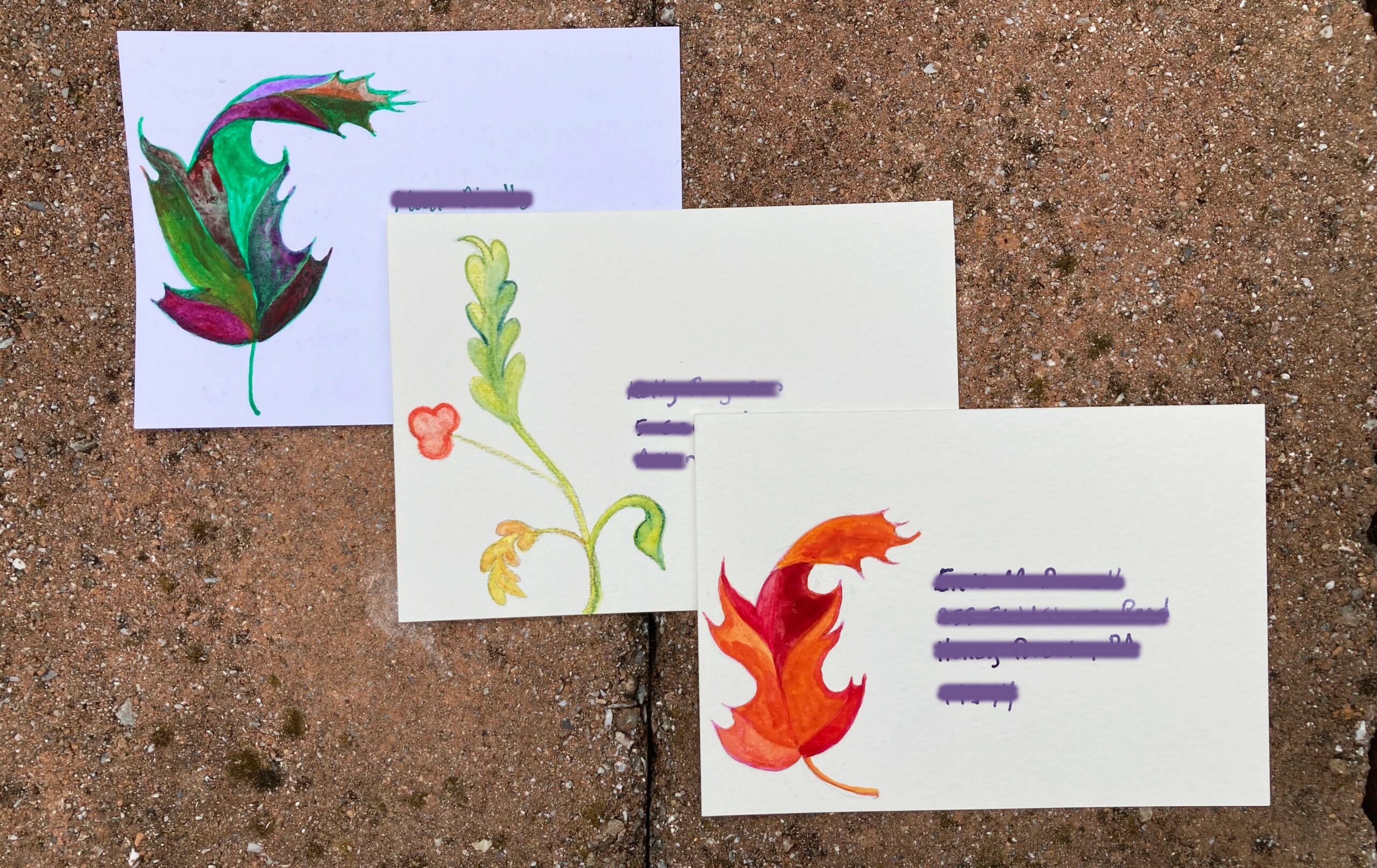 Three postcards on a brick wall, address side up, in a cascade from top left to bottom right. The first has a fall oak leaf with disparate colors (green, dark crimson, gold, purple), the second a stylized floral motif, and the third another leaf, this time in gold, orange, and crimson. The recipient information is obscured.