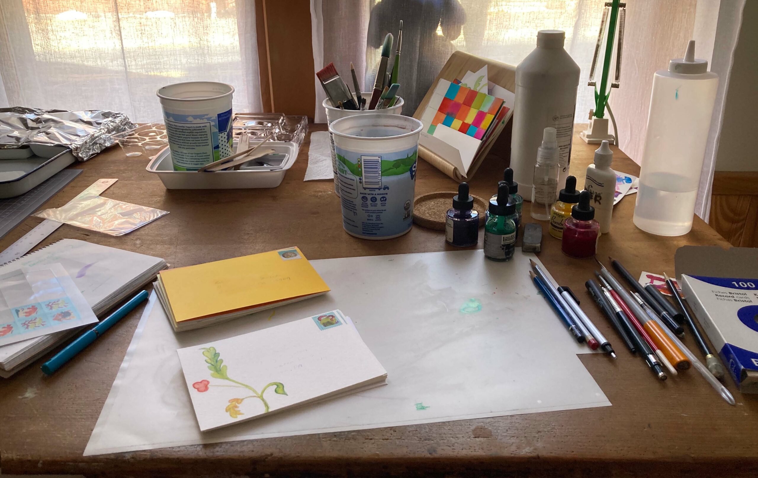 Work table with two stacks of postcards, stamp sheet, paintbrushes, drawing tools, ink bottles, and a jug of gesso. The cards have been addressed but the information is obscured.